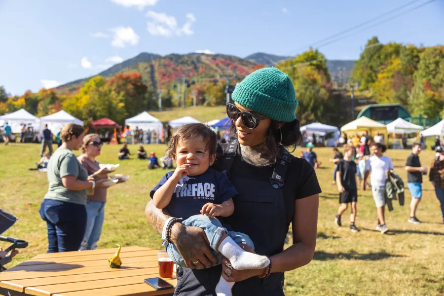 A smiling woman holds a baby in front of a mountainside dotted with people and tents at a festival.